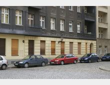 Straßenszene mit einer Reihe geparkter Autos vor einem mehrstöckigen Wohngebäude mit dunkelgrauer und gelber Fassade, geschlossenen Fensterläden und dekorativen Elementen. Der Gehweg und die gepflasterte Straße erstrecken sich im Vordergrund.