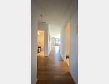 Flur in einer modernen Wohnung mit hellem Holzfußboden, weißen Wänden und Türen zu angrenzenden Räumen. Am Ende des Flurs ist ein helles Fenster mit Vorhängen und Ausblick sichtbar.