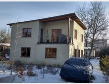 Zweistöckiges Wohnhaus mit hell verputzter Fassade, großen Holzrahmenfenstern und einem kleinen Balkon mit Metallgeländer. Im Vordergrund befindet sich ein mit Schnee bedeckter Garten und ein unter einer Plane abgedecktes Auto. Im Hintergrund stehen unbelaubte Bäume.