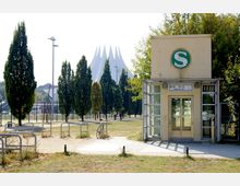 Eingang zu einer S-Bahn-Station mit einem „S“-Logo auf einem rechteckigen Gebäude, umgeben von Bäumen und einem Fahrradständer im Vordergrund. Im Hintergrund befindet sich das weiße Dach des Berliner Tempodroms, erkennbar an seinen spitzen, zeltartigen Strukturen.