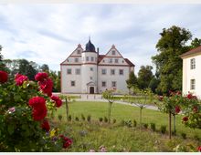 Barockes Herrenhaus mit weißer Fassade, roten Fensterrahmen und einem Turm mit schwarzer Kuppel, umgeben von gepflegtem Garten mit Rosensträuchern und grünen Bäumen im Hintergrund.