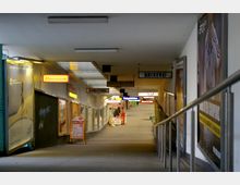 Einkaufspassage mit schmalem Gang und Geschäften auf beiden Seiten, darunter eine Bäckerei und eine Konditorei, erkennbar an Leuchtschildern. Die Passage ist überdacht, mit Werbeplakaten an den Wänden und einer leicht ansteigenden Rampe.