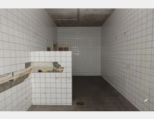 Raum mit weißen Fliesen an den Wänden und einem grauen Boden aus Fliesen. Eine beschädigte Wandpartie ist sichtbar, der Raum wirkt leer und weist eine schlichte Architektur mit einer Tür im Hintergrund auf.