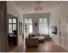 Helles Wohnzimmer in einem Altbau mit weißen Wänden, Stuckelementen an der Decke und großen Fenstern mit transparenten Gardinen. Der Raum ist mit einem beigen Sofa, einem an der Wand montierten Fernseher und Holzdielen versehen; im Hintergrund ist ein kleiner Arbeitsbereich mit Schreibtisch und Stuhl zu sehen.