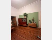 Wohnraum mit Holzdielenboden, einer hellgrünen Wand und weißen Wänden. Ein Holzschrank und eine Kommode stehen an der Wand, darauf Pflanzen und Dekorationsgegenstände; eine Tür und ein kleiner Teil eines Holzstuhls sind ebenfalls sichtbar.