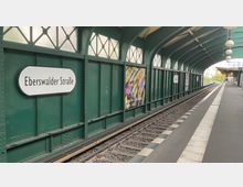Ein oberirdischer U-Bahnhof mit dem Namen „Eberswalder Straße“, gekennzeichnet durch grüne, metallene Wände mit geschwungenen Fensterelementen und ein gewölbtes Dach. Die Gleise verlaufen parallel zu einem breiten Bahnsteig mit Lampen und Informationsanzeigen.