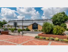 Ein modernes Gebäude mit der Aufschrift „Havelauenhalle“ auf der Frontseite, errichtet aus gelben Klinkersteinen und einem geneigten Metalldach. Der Eingangsbereich ist von Grünflächen und Wegen mit rotem Pflaster umgeben, im Hintergrund sind Bäume und ein teilweise bewölkter Himmel zu sehen.