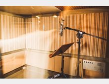 Tonstudio-Aufnahmeraum mit schallabsorbierenden, gestreiften Wänden. Im Vordergrund ein Mikrofon an einem Galgenstativ und ein Notenpult, umgeben von technischer Audio-Ausstattung.