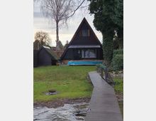 Dunkelbraunes A-Frame-Haus mit einem Spitzdach, gelegen an einer Rasenfläche mit Blick auf ein Gewässer. Im Vordergrund führt ein schmaler Holzsteg über das Wasser, umgeben von Bäumen und Sträuchern.