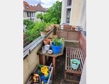 A small balcony with wooden flooring and railings, featuring potted plants, gardening tools, and a wooden bench. The setting overlooks a residential area with trees and pitched-roof houses in the background.