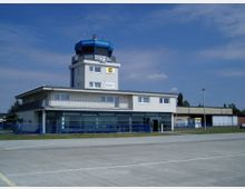 Kleiner Flughafen mit einem Kontrollturm und einem einstöckigen Gebäude in modernem Design. Im Vordergrund ein betonierter Vorplatz, im Hintergrund blaues Geländer und umliegende Hangars.