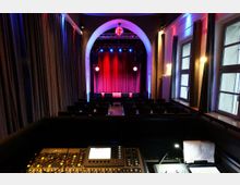 Blick aus dem Technikbereich in einen kleinen Theatersaal mit Sitzreihen und einer Bühne mit rotem Vorhang unter einem großen Rundbogen. Rechts sind hohe Fenster und Wandlampen zu sehen, die den Raum in farbigem Bühnenlicht beleuchten.