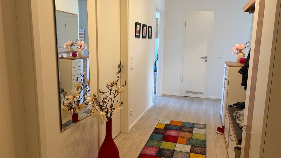 Heller, moderner Wohnungsflur mit hellem Holzboden, weißen Wänden und mehreren Türen entlang des Gangs. An der Wand hängen ein großer Spiegel und kleine Bilder; auf dem Boden liegt ein bunter Patchwork-Teppich, daneben steht eine rote Bodenvase mit künstlichen Blumen und rechts befindet sich eine Garderobe mit Kommode und Holzablage.