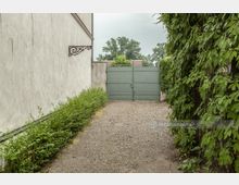 Schmaler, mit Kies bedeckter Weg zwischen einer hellen, verputzten Hausfassade links und einer mit grünem Efeu bewachsenen Wand rechts, der zu einem geschlossenen, grün gestrichenen Eisentor führt. Im Hintergrund sind Bäume und Dächer sichtbar.