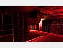 Ein dunkler, industrieller Raum mit Betonwänden und -decke, beleuchtet von rotem Licht. Der Raum enthält zahlreiche offene Regalstrukturen, teilweise mit Graffiti versehen, und eine zentrale Säule mit Belüftungsrohren.