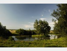 Landschaft mit einem ruhigen Fluss, umgeben von grünen Wiesen und Bäumen. Im Hintergrund befinden sich ein Feld mit Heuballen und ein Traktor unter einem klaren Himmel.