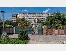 Mehrstöckiges Wohngebäude mit Balkonen und bunten Sonnenschirmen im Hintergrund, umgeben von einem gepflegten Garten mit Bäumen, Sträuchern und einem kleinen Tor aus Metall und Holz im Vordergrund. Der Garten ist von Zäunen begrenzt, während ein gepflasterter Weg davor verläuft.