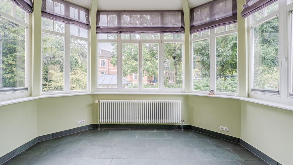 Heller, leerer Erkerraum mit großen, umlaufenden Sprossenfenstern und halbtransparenten Raffrollos. Die Wände sind hellgrün, am Fenster steht ein weißer Heizkörper, und der Boden ist mit dunkelgrauen Fliesen ausgelegt; draußen sind Bäume und Nachbarhäuser zu sehen.