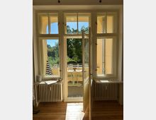 Ein sonnendurchfluteter Innenraum mit Holzfußboden und großen, weißen Doppelfenstern, die zu einem kleinen Balkon führen. Auf dem Balkon stehen ein Tisch und zwei Stühle, umgeben von Geländern, mit Blick auf grüne Bäume.