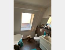 Modernes Badezimmer mit einer freistehenden weißen Badewanne, einem Dachfenster, einer wandhängenden Toilette und einem Waschtisch mit großem Spiegel. Der Raum hat braune Fliesenböden, helle Wände und eine Dekoration mit Pflegeprodukten und einem kleinen hölzernen Beistelltisch.