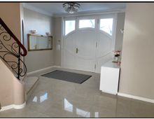 Eingangsbereich eines Hauses mit großen weißen Doppeltüren, einer Treppe mit schmiedeeisernem Geländer und glänzendem Fliesenboden. An der Wand ein goldgerahmter Spiegel, seitlich ein weißer Schrank mit Blumenvasen.