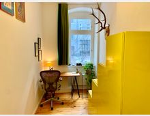 Kleines Arbeitszimmer mit Holzboden, einem Schreibtisch vor einem Fenster mit grünem Vorhang, gelbem Regal an der rechten Wand und Wanddekorationen wie Bilderrahmen und einem Hirschgeweih. Pflanzen auf dem Fensterbrett und Tisch ergänzen die Einrichtung.