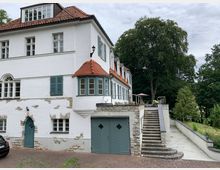 Weißes zweistöckiges Gebäude mit roten Ziegeldächern, blauen Fensterläden und Natursteinelementen an der Fassade. Eine Außentreppe führt zu einer erhöhten Terrasse mit Geländer und Möbeln, umgeben von Bäumen und einer gepflasterten Fläche.