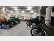 Ein Ausstellungsraum in einem Automuseum zeigt zwei antike Autos mit grünen Karosserien, goldenen Details und Holzrädern. Im Hintergrund sind historische Poster, Motorräder und weitere Ausstellungsstücke zu sehen.