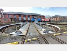 Drehscheibe einer historischen Eisenbahnwerkstatt mit Schienenführung und Geländern, im Hintergrund Backsteingebäude und abgestellte Lokomotiven. Die Umgebung zeigt Bewuchs und Bahninfrastruktur.