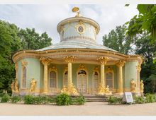 Rokokopavillon mit runder Grundform und grünlicher Fassade, verziert mit vergoldeten Statuen, Säulen und Dekorationen. Das Gebäude steht in einer Gartenanlage, umgeben von Bäumen und Hecken.