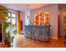 Bright, modern home bar area with a curved blue speckled counter, clear acrylic stools, and three arched backlit bottle shelves mounted on a pale wall. Large windows with mustard-yellow curtains, hanging pendant lights, indoor plants, and wood plank flooring define the space.