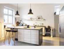 Moderne Küche mit einer Kücheninsel aus Holzlaminat und zwei Barhockern. Helle Wände, ein großes Fenster, Hängelampen, offene Regale mit Geschirr sowie ein Edelstahlkühlschrank und Kaffeemaschine prägen den Raum.