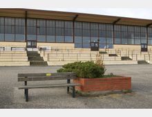 Ein leerer Vorplatz mit einer Holzbank und einem Pflanzkasten aus roten Ziegelsteinen im Vordergrund. Im Hintergrund steht ein Gebäude mit großen Fenstern und einer überdachten Tribüne mit Geländern und Stufen.