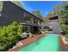 Modernes Wohnhaus mit dunkler Holzfassade, großen Glasfenstern und einer umlaufenden Terrasse. Im Vordergrund befindet sich ein länglicher Swimmingpool, umgeben von Pflanzen und Sitzgelegenheiten, eingebettet in eine grüne Umgebung mit Bäumen.
