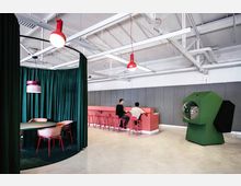 Ein bunter, moderner Büroraum mit einer Sitzecke hinter einem dunkelgrünen Vorhang, einem roten Tresen mit hohen Hockern, an dem zwei Personen sitzen, und einer grünen, futuristischen Kabine mit einer transparenten Glaskuppel. Die Decke ist industriell gestaltet mit sichtbaren Rohren und Lampen.