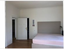 Schlafzimmer mit einem großen Bett ohne Bettwäsche, weiß gestrichenen Wänden und Holzboden. Links steht eine geöffnete weiße Tür, die zu einem angrenzenden Raum führt.