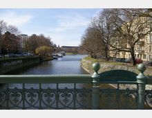 Blick von der Admiralsbrücke auf einen breiten Flusskanal, gesäumt von Bäumen und historischen Gebäuden auf der rechten Seite sowie moderneren Bauten auf der linken Seite. Boote sind am Ufer festgemacht, und die Szenerie zeigt eine friedliche städtische Atmosphäre.