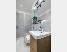 Modernes Badezimmer mit grauen Steinfliesen, bodengleicher Dusche mit Glasabtrennung und Waschtisch mit weißem Aufsatzbecken auf Holzunterschrank. Integrierte Ablage mit Handtüchern und Pflegeprodukten sowie Decken-Spots und Schienenbeleuchtung.