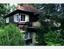 Zweistöckiges Holzhaus mit dunkler Fassade, weißen Fensterrahmen und gelben Fensterläden, umgeben von dichter Vegetation. Das Gebäude hat ein schräges Dach mit roten Dachziegeln und einen Balkon im Obergeschoss.