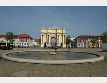 Ein Platz mit einem runden Brunnen im Vordergrund, dahinter das barocke Brandenburger Tor in Potsdam, flankiert von historischen Häusern mit roten Dächern. Im Hintergrund sind Bäume und ein klarer Himmel sichtbar.