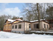 Gebäude einer Waldgaststätte in ländlicher Umgebung mit einer Fassade aus gelbem Putz und roten Backsteinelementen, ergänzt durch einen modernen Anbau mit großen Fenstern. Im Vordergrund liegt eine verschneite Landschaft, umgeben von kahlen Bäumen.