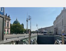 Blick über eine Berliner Straße mit dem Fernsehturm im Hintergrund, der Berliner Dom links und historische Gebäude rechts. Im Vordergrund befindet sich der Eingang zu einer U-Bahn-Station mit Rolltreppen.