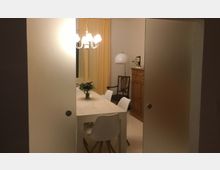 Esszimmer mit einem rechteckigen weißen Tisch, umgeben von weißen Stühlen. Im Hintergrund sind ein Holzschrank, ein Polsterstuhl, eine Stehlampe und gelbe Vorhänge zu sehen, beleuchtet von einem hängenden Kronleuchter.