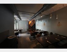 Innenansicht eines modernen Restaurants mit langen dunklen Tischen und Stühlen in einem schmalen Raum. Sichtbare Lüftungsrohre an der Decke, hängende Pendelleuchten und graue Wände prägen das industrielle Ambiente.