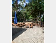Außenbereich eines Cafés oder Restaurants mit Holztischen und -bänken auf einem gepflasterten Boden, umgeben von hohen Bäumen. Im Hintergrund befindet sich ein umzäunter Spielplatz mit Spielgeräten, daneben steht eine Tafel mit einem Hinweistext.
