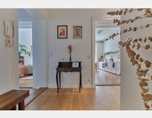 Wohnungsflur mit Holzboden, in dessen Mitte ein antiker Beistelltisch mit dekorativer Vase und gerahmten Bildern steht. Links führt eine Tür in ein Wohnzimmer mit Pflanzen und einem Sideboard, rechts eine Tür in eine Küche mit weißen Schränken und einer hohen Decke mit Stuckverzierungen.