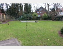 Weitläufiger Garten mit einer Rasenfläche, vereinzelten Sträuchern und Bäumen sowie einer gepflasterten Fläche im Hintergrund. Im Hintergrund sind ein rotes Backsteinhaus und dichte Hecken zu sehen, während im Vordergrund ein Holzdeck sichtbar ist.