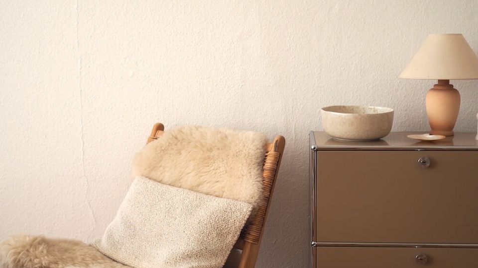 Cozy indoor corner with a wicker lounge chair draped in cream sheepskin and a blanket on a wooden floor, beside a small wooden stool. A low metal cabinet against a white textured wall holds a stone bowl and a table lamp.