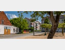 Wohngebiet mit einer Kombination aus alten und neuen Gebäuden, darunter ein weißes Haus mit rotem Ziegeldach und ein Mehrfamilienhaus mit Balkonen. Im Vordergrund sind ein gepflasterter Platz, kleine Bäume und ein Baumstamm sichtbar, während im Hintergrund ein blauer Himmel mit vereinzelten Wolken zu sehen ist.