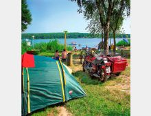 Ein Campingplatz am Ufer eines Sees mit einem grünen Zelt, einer roten Motorradgespann und mehreren Personen, die auf einer Wiese sitzen oder im Wasser schwimmen. Im Hintergrund ist dicht bewaldetes Ufer sichtbar.